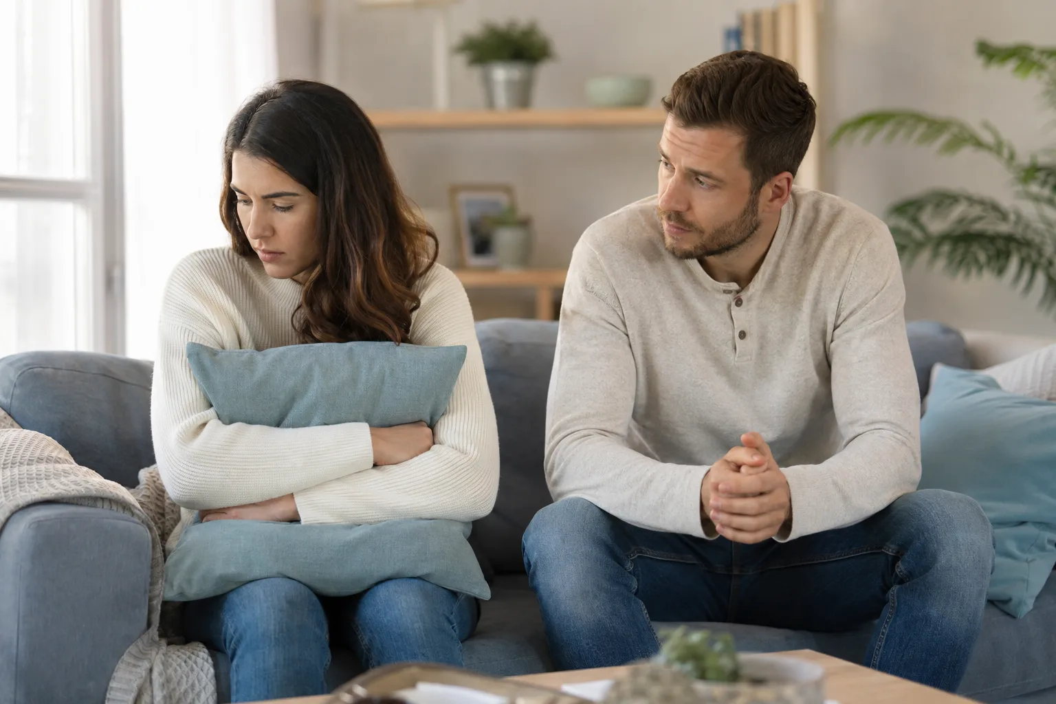 An upset woman holds a cushion while her concerned partner sits nearby unsure how to comfort her